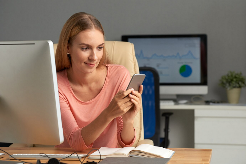 Женщина сидит за столом с компьютером и смотрит на свой смартфон.